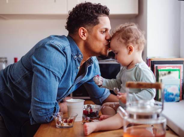 A father and a small toddler son eating fruit and yoghurt indoors at home. A father and a small toddler son eating fruit and yoghurt in kitchen indoors at home. iberian ethnicity stock pictures, royalty-free photos & images