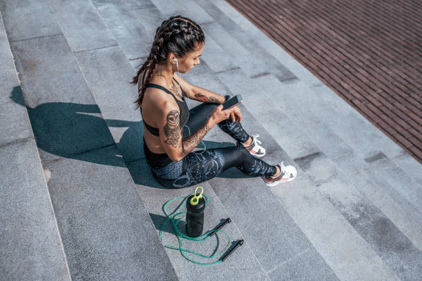 Girl athlete sitting steps resting after workout fitness summer city, day morning. Sportswear, smart phone, headphones, jumper. Active lifestyle, motivation power. Internet application, music. stock photo