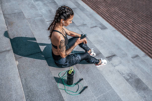 girl athlete sitting steps resting after workout fitness summer city, day morning. sportswear, smart phone, headphones, jumper. active lifestyle, motivation power. internet application, music. - headphones women tattoo music imagens e fotografias de stock