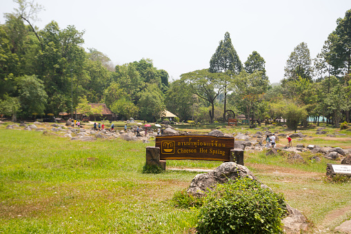 Area of hot springs of Chae Son national park in Lampang province. In front of springs is a wooden sign with text. People are at springs for boiling eggs in baskets in water.