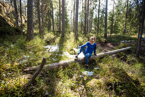 Female holding mobile phone and a bottle of water in evergreen forest.