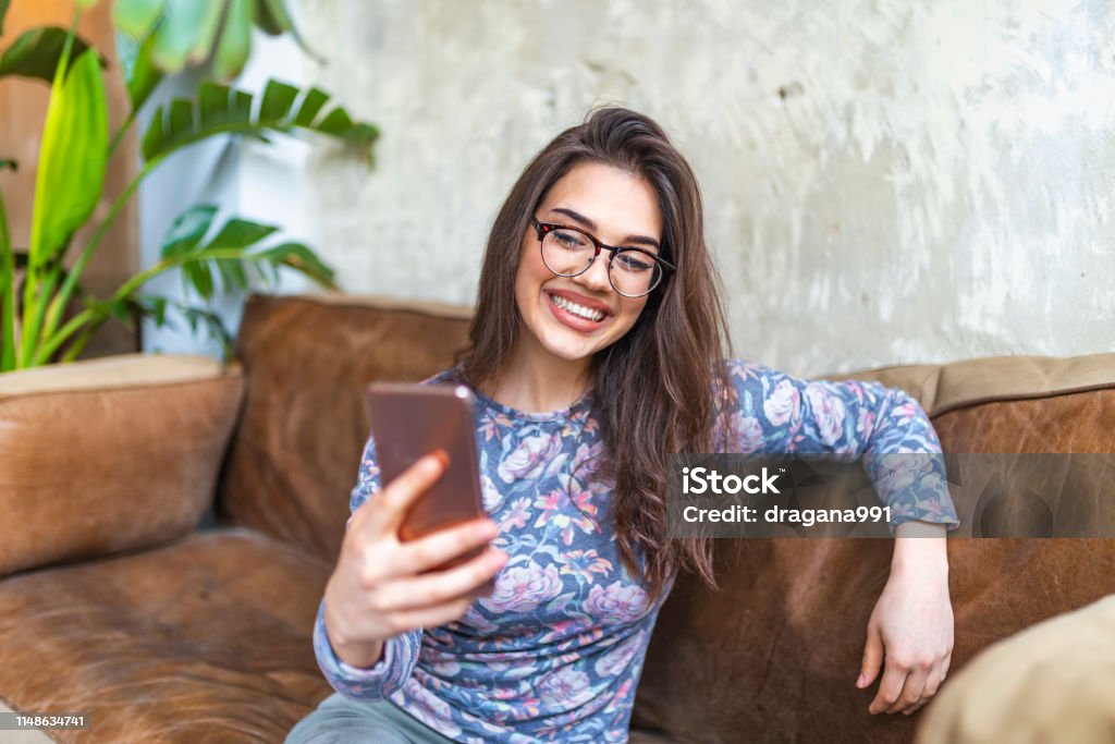 Happy young woman holding mobile phone on hands and reading message at home Happy young woman holding mobile phone on hands and reading message at home, Smiling hipster girl chatting with friends while seating in sunny cafe shop 20-29 Years Stock Photo