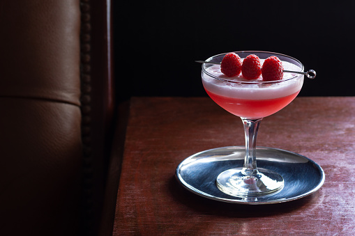 A Clover Club cocktail in a couple glass on a dark wood table next to a leather seat. This drink is made from gin, lemon juice, raspberries, and vermouth shaken with egg white to give it a foamy layer on top. Taken in a dark luxurious bar or restaurant.
