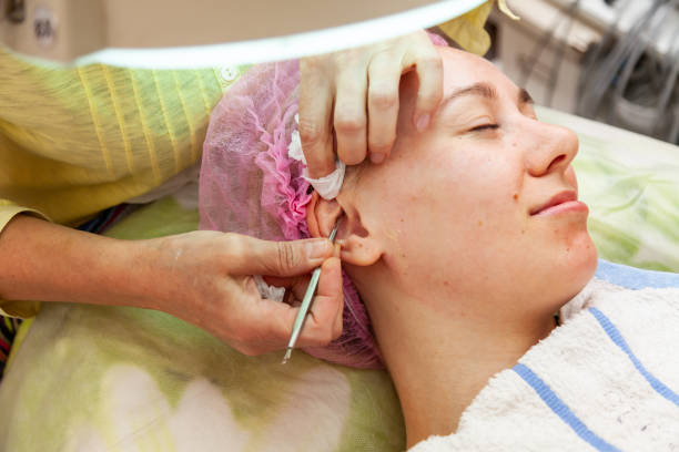 A young girl is lying on a couch during cosmetic procedures with a mask on the face above which beautician woman squeezes body fat and pimples with special metal tool in the form of a blade in ear una niña está acostada en un sofá durante los procedimientos cosméticos con una máscara en la cara por encima de la cual la mujer esteticista aprieta grasa corporal y granos con una herramienta especial de metal en forma de una cuchilla en el oído