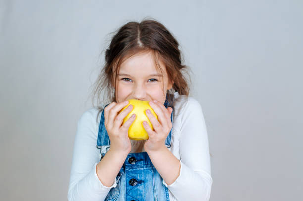 portrait émotionnel petite belle fille avec des tresses en jeans salopette mangeant des piqûres tenant une pomme. 6-7 ans studio - 6 7 years little girls child standing photos et images de collection