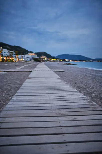 wooden footpath on a rocky beach sea on the right side and hotel on the left side, cloudy evening and street lights that illuminate