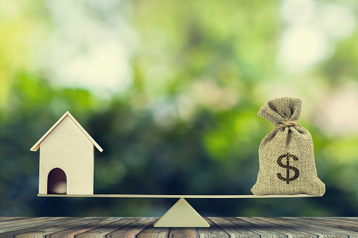 Money and home,loan,mortgage. Change home into cash concept. US Dollar in sack bag, Wooden house model put on scales on wood table with green tree bokeh as background. Balance home and debt.