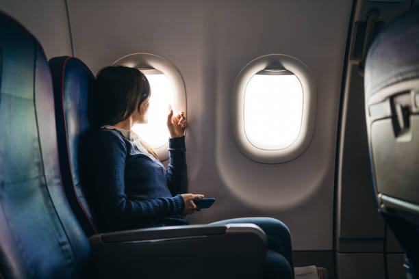 Looking through airplane window Young woman sitting in an airplane, waiting for a flight to start, looking through airplane window, holding mobile phone. window seat vehicle stock pictures, royalty-free photos & images