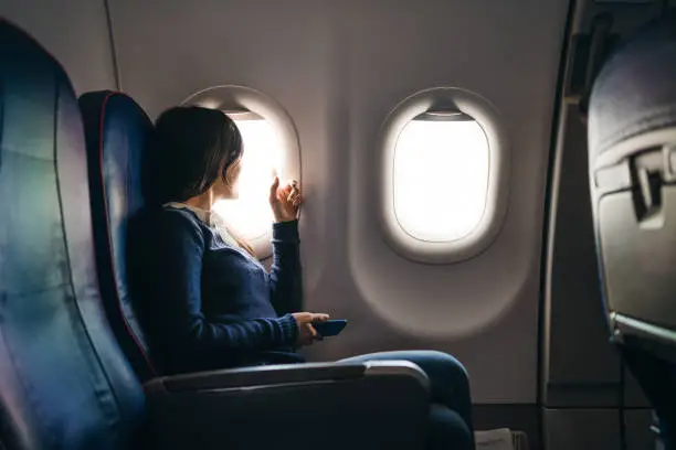 Young woman sitting in an airplane, waiting for a flight to start, looking through airplane window, holding mobile phone.