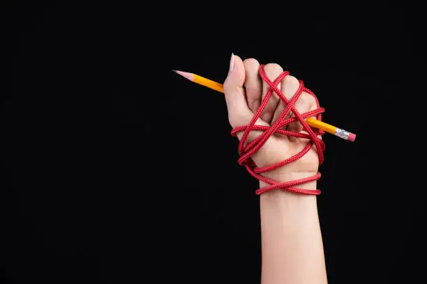 Photo of Woman hand with yellow pencil tied with red rope, depicting the idea of freedom of the press or expression on dark background. World press freedom and international human rights day concept.