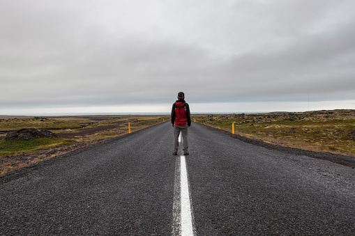 Snaefellsness, Iceland - July 22, 2016. Man standing on Iceland road leading straight to the horizon. Straight road line in rural Icelandic landscape on a cloudy summer day.