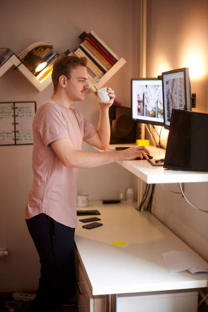 one young man, standing at standing desk, working with his computer at his home, in his room. one young man, 20-29 years old, standing at standing desk, working with his computer at his home, in his room. standing desk photos stock pictures, royalty-free photos & images