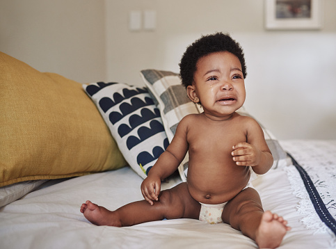 Shot of an adorable baby crying while sitting on a bed at home