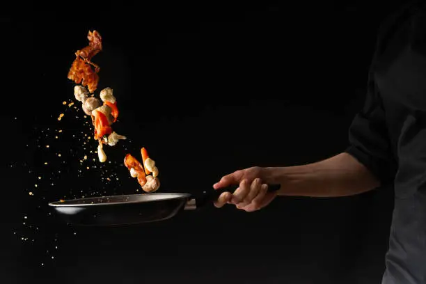 Cook prepares bacon pieces with garlic and hot pepper in a pan, freeze in the air, on a black background, recipe-book, menu, gastronomy, culinary background