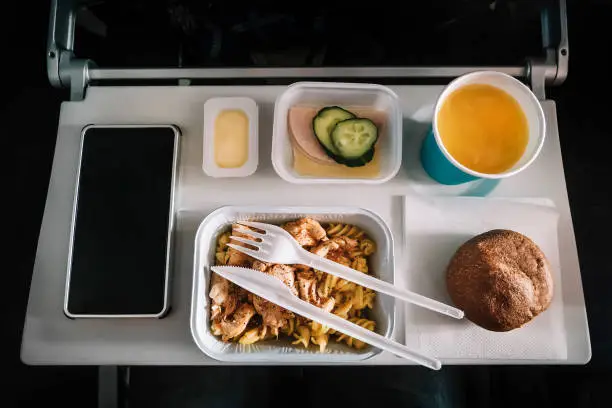 Inflight meal service tray for economy class, meat with pasta, seasoning fruit, salad, cucumber, a glass of juice and butter. selective focus.