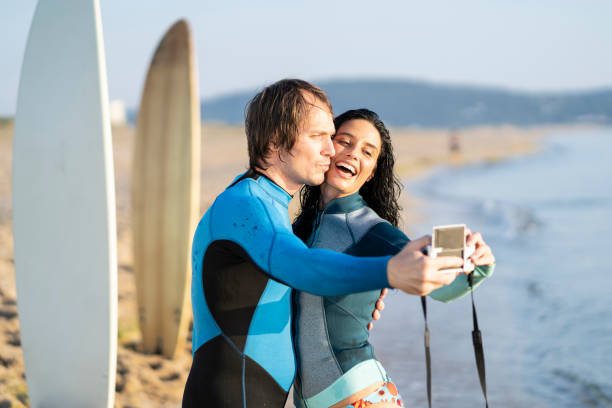 istock Happy couple taking selfie with camera on beach, surfers 1148782140