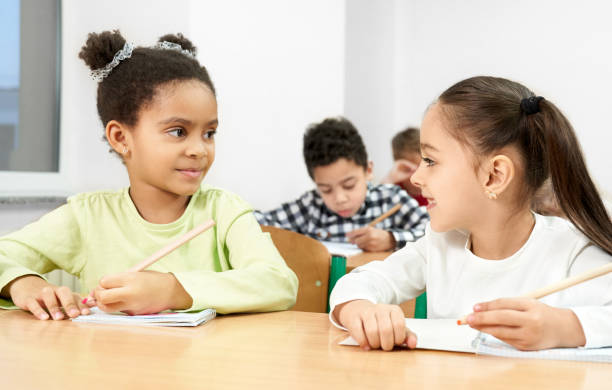 istock Girls at desk in classroom, looking at each other, posing. 1148791328