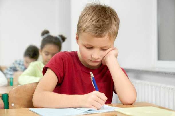 istock Schoolboy at desk writing with pen in copybook. 1148791347