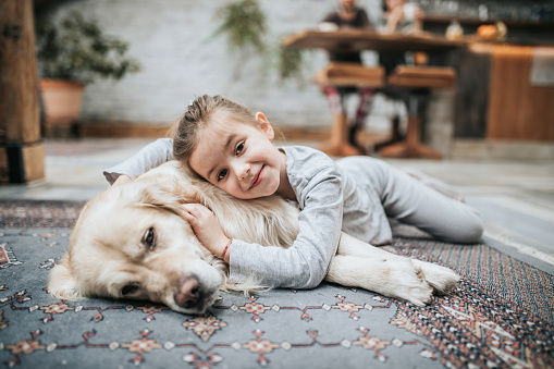 Cute little girl relaxing with her dog on carpet at home and looking at camera.