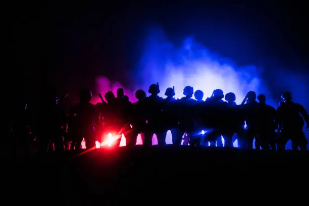 Photo of Anti-riot police give signal to be ready. Government power concept. Police in action. Smoke on a dark background with lights. Blue red flashing sirens.