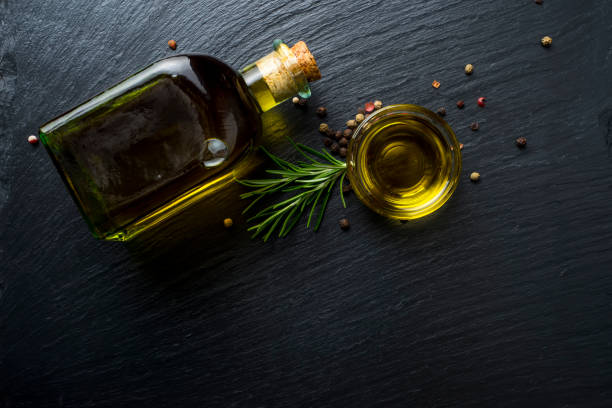 virgin olive oil in a glass bottle, rosemary and peppercorns on black stone slate - virgin olive oil imagens e fotografias de stock