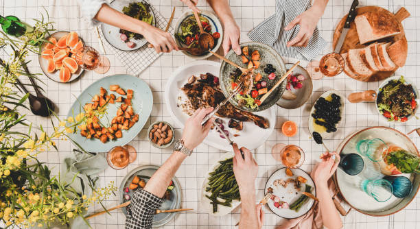 Peoples hands with various snacks and blooming mimosa, top view Family or friends gathering dinner. Flat-lay of peoples hands, roasted lamb shoulder, salads, vegetables, rose wine, mimosa branches over white checkered tablecloth, top view. Celebration party dinner food flat lay stock pictures, royalty-free photos & images