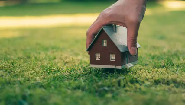 Photo of Concept of selling a house. A hand is holding a model house above green meadow.