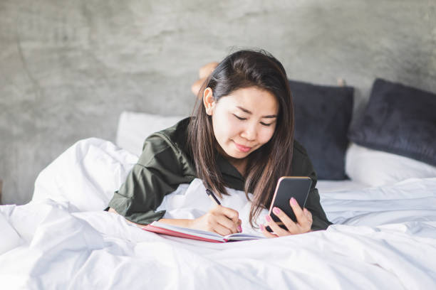 istock Asian woman lying down on bed hand writing on notebook hand holding smart phone 1148815233