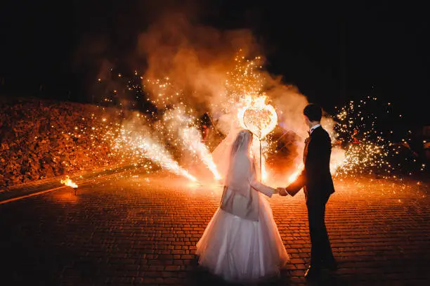 Wedding Fire show and fire heart at night on black background.
Burning heart with fireworks. Brides holding hands and looking at fireworks and fire show.
