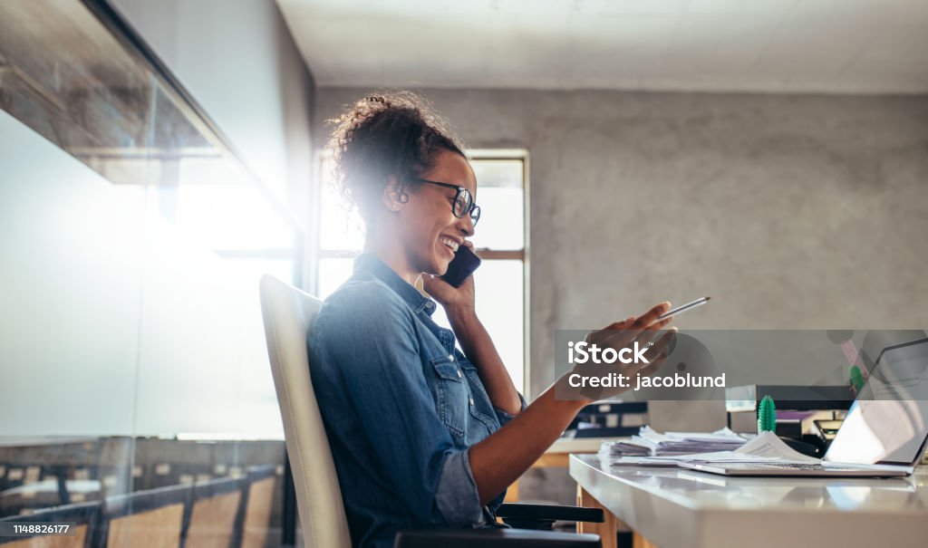 Businesswoman in conversation over phone Smiling young woman talking on phone with a laptop in front. Businesswoman in conversation over phone at office. Owner Stock Photo