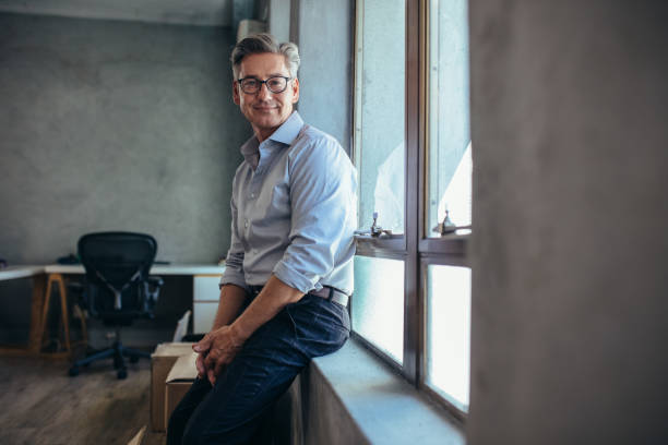 Entreprenuer at his office Happy mature businessman leaning by a window sill and staring to camera. Male entrepreneur at his office looking at camera. mature businessman stock pictures, royalty-free photos & images