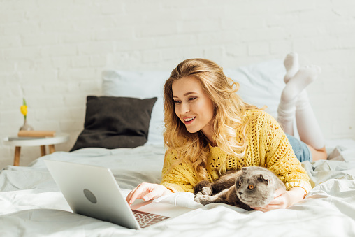 beautiful smiling girl lying in bed with scottish fold cat and using laptop at home