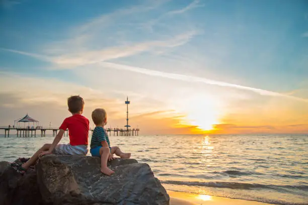 Photo of Two boys sitting on the rock at the beach at sunset
