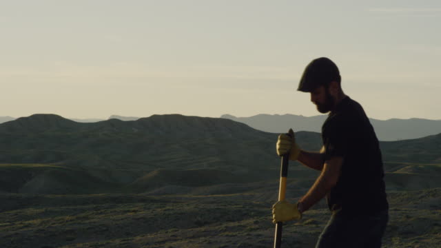 A Caucasian Man in His Thirties with a Beard and Wearing a Hat and Work Gloves Digs a Hole in the Ground with a Shovel in the Desert at Sunset
