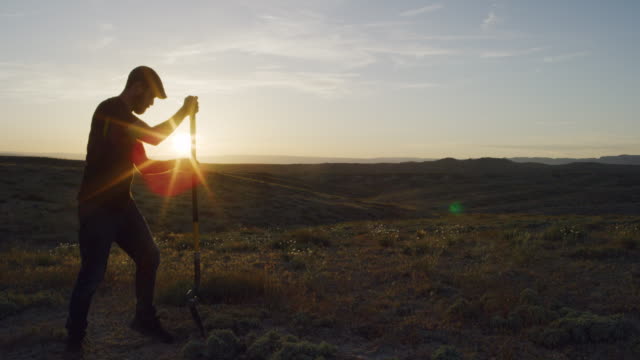 A Caucasian Man in His Thirties Wearing a Hat Digs a Hole in the Ground with a Shovel in the Desert at Sunset