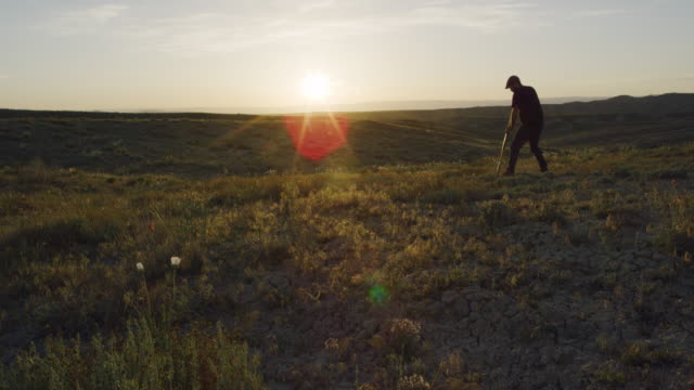 A Caucasian Man in His Thirties Wearing a Hat and Work Gloves Digs a Hole in the Ground with a Shovel in the Desert at Sunset