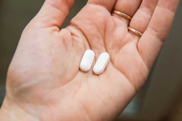 Close-up view of a hand holding two white pills in the palm above a blurry background, painkiller stock photo