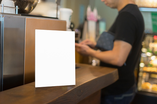 Mock up Menu frame on Table in Bar restaurant or coffee shop ,Stand for booklets with white sheets of paper acrylic tent card on cafeteria ,barista preparing cup of coffee for customer in background.