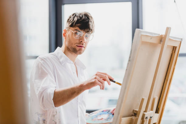 istock selective focus of handsome artist in glasses looking at camera while drawing on canvas 1148855923