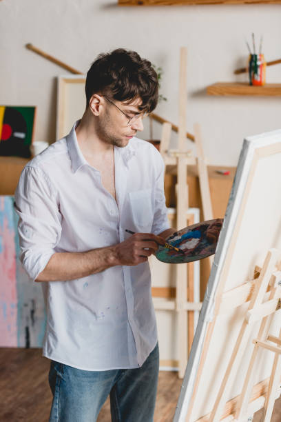istock handsome artist mixing paints on palette while standing near easel with canvas 1148855925