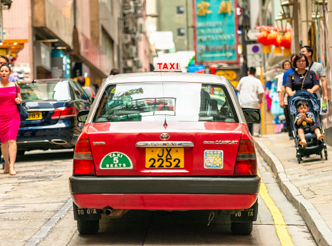 Hong Kong Island, Hong Kong SAR - A rear view of a taxi cab passing pedestrians on a narrow Hong Kong street, with its light on looking for a passenger.