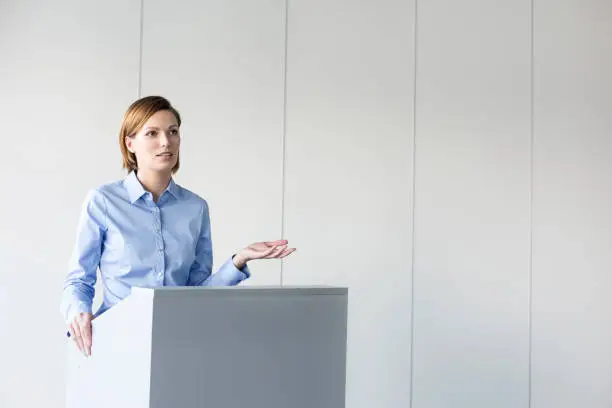 Photo of Confident businesswoman giving speech at podium against wall in office