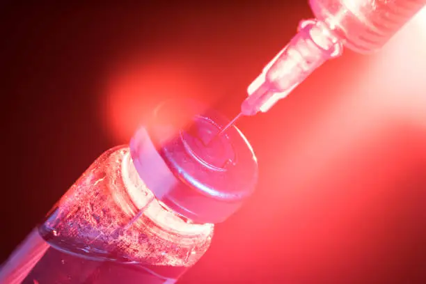 A glass medical bottle with a vaccine and a disposable syringe in color light. A conceptual photo about the danger of viral diseases