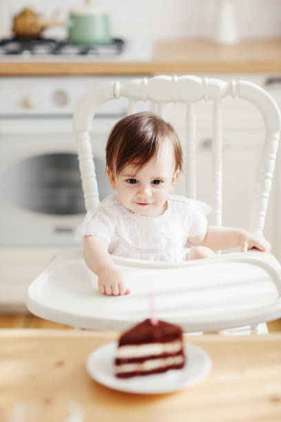 かわいい女の赤ちゃんは、ハイチェアに座って、キャンドルとバースデーケーキの一部を見て、彼女の小さな手でそれに到達しようとしています - cake birthday domestic kitchen child ストックフォトと画像