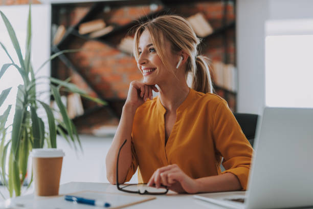Cheerful woman wearing wireless earphones and smiling Listening to music. Happy young woman sitting in the office with laptop and coffee while using modern wireless earphones blouse stock pictures, royalty-free photos & images
