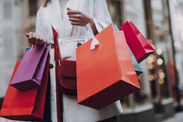 Elegant young woman with shopping bags standing on the street Close up of lady in white trench coat holding colorful shopping bags merchandise stock pictures, royalty-free photos & images