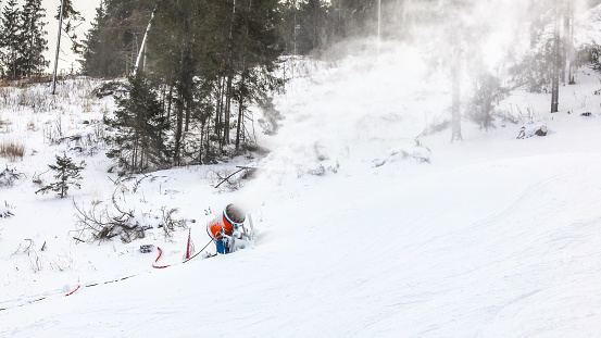 Snowgun spray artificial ice crystals over ski piste, snowmaking in winter sports resort, trees in background