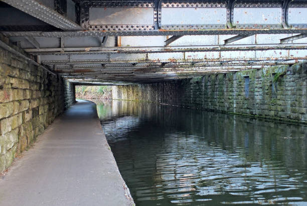 blick unter einem alten low-steel-träger-bräghaus, der die leeds zum liverpool kanal in der nähe einer armlege mit steinmauer und einem schmalen fußweg entlang des wassers überquert - aredale stock-fotos und bilder