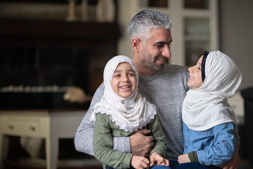 A young ethnic girl is laughing and smiling while interacting with her Muslim dad and sister in their living room.