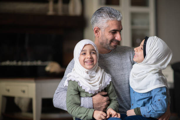 A Young Muslim Girl Enjoying Cuddle Time With Her Dad and Sister A young ethnic girl is laughing and smiling while interacting with her Muslim dad and sister in their living room. pics of a cute muslim outfits stock pictures, royalty-free photos & images